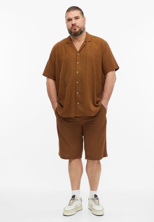 Mann mit Bart, der ein braunes kurzärmeliges Hemd mit Knöpfen und passende Shorts, weiße Socken und beige Turnschuhe trägt, steht mit den Händen in den Taschen.