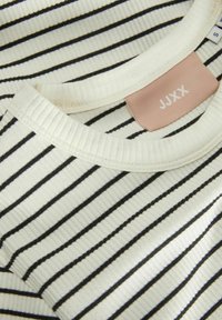 Chemise rayée blanche avec de fines lignes noires, présentant une texture côtelée et un col rond. Étiquette "JJXX" en rose clair.