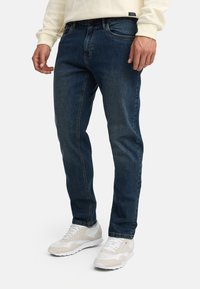 Man die donkere blauwe jeans, witte sneakers en een crème kleurige gebreide trui draagt, staand tegen een effen lichte achtergrond.