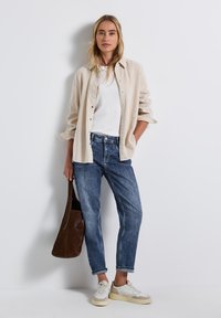 Lichtbeige corduroy shirt over een wit T-shirt, gecombineerd met blauwe jeans en beige sneakers. Een bruine tote bag hangt over één schouder.