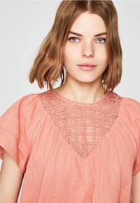 Blusa color pesco con maniche corte e fluide e un motivo in pizzo floreale sullo scollo, realizzata in tessuto leggero e strutturato.