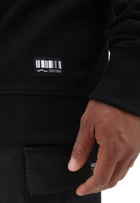 Felpa nera con etichetta a codice a barre. Primo piano del tessuto testurizzato e di una mano di una persona appoggiata vicino a una tasca laterale.