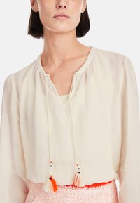 Cremfarbene Bluse mit gerafftem Ausschnitt und Quasten-Details. Verfügt über einen V-Ausschnitt und einen leichten, durchsichtigen Stoff.