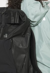 Black waterproof backpack with a roll-top design and logo. Light green hooded jacket with a sleek texture and logo on the back.