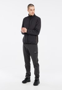 Sort zip-up jakke med høj krave, glat tekstur; parret med mørke bukser med en lynlås lomme, suppleret med sorte sko.