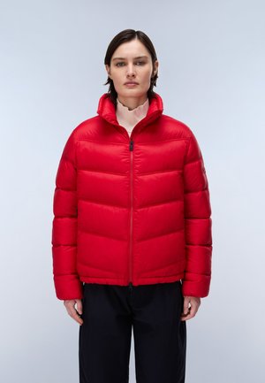 Donna con capelli corti che indossa una giacca imbottita rossa brillante sopra un dolcevita chiaro e pantaloni neri, in piedi davanti a uno sfondo chiaro uniforme.