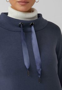 Marineblauer Pullover mit hohem Kragen und silbernen Ösen. Verfügt über Satinschnüre mit metallischen Enden und einem strukturierten weißen Rollkragen darunter.