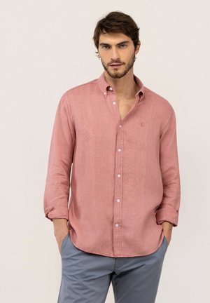 Hombre con cabello castaño y barba, vistiendo una camisa rosa de manga larga con botones y pantalones grises, con las manos en los bolsillos, sobre un fondo liso.