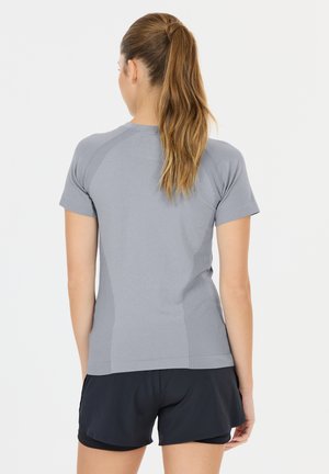 Kvinde med lang brunt hestehale iført en tætsiddende grå kortærmet skjorte og sorte atletikshorts, stående med ryggen mod på en ensfarvet baggrund.