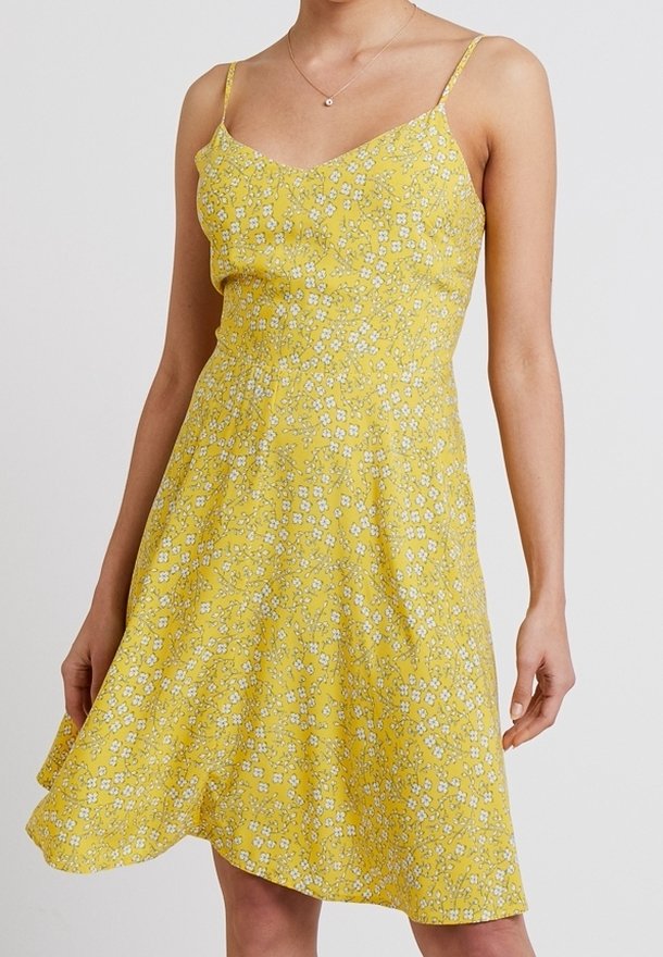 Gul blommig klänning med tunna axelband, med en figursydd överdel och utsvängd kjol, prydd med vita och gröna blommönster. Mjuk tygtextur.