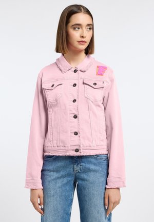 Junge Frau schaut nach rechts, trägt eine hellrosa Jeansjacke mit Knopfleiste und blaue Jeans vor schlichter Hintergrund.