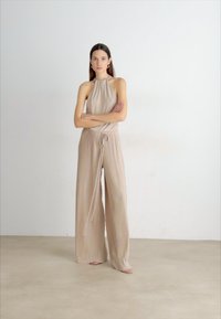 Beige, ribbed jumpsuit with a halter neck design, adjustable drawstring waist, and wide-leg silhouette, creating a relaxed fit.