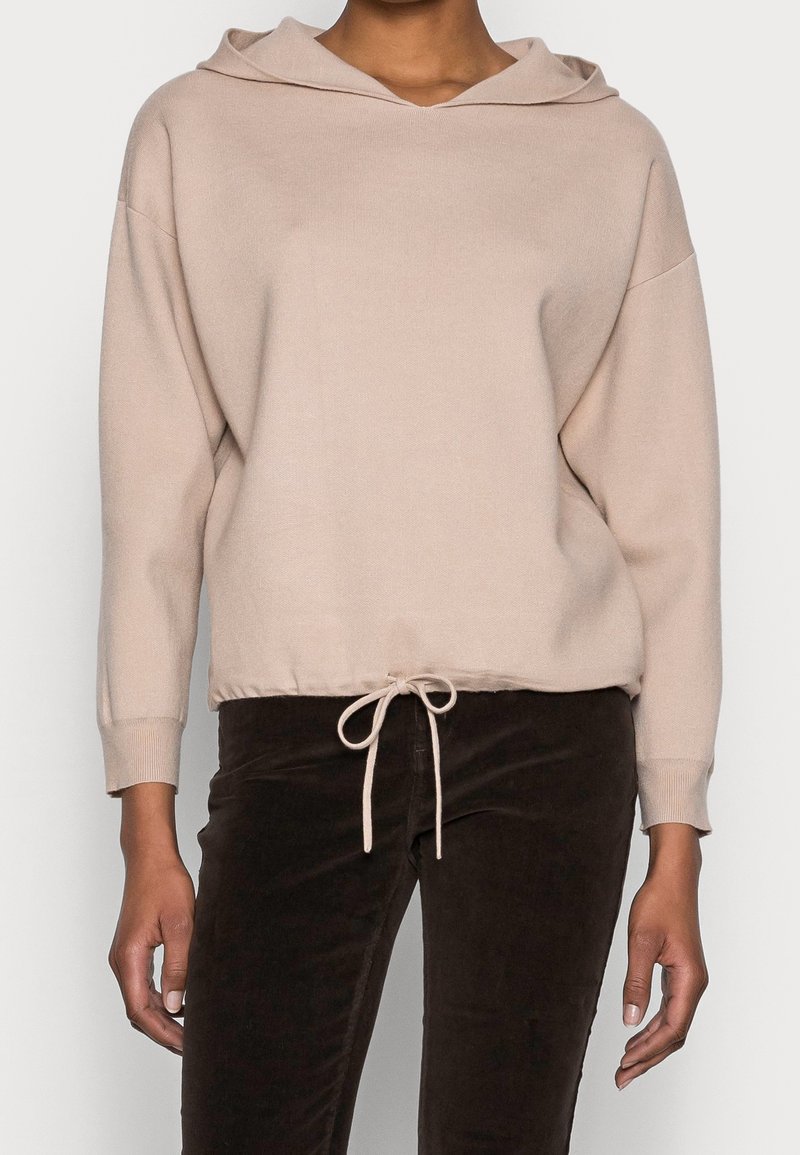 Sweat à capuche beige à manches longues avec taille à cordon, porté avec un pantalon marron foncé, sur une personne vue du cou jusqu'aux cuisses.