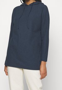 Persona che indossa un hoodie lungo a maniche lunghe di colore blu navy con cordini e pantaloni di colore bianco sporco, in piedi contro uno sfondo neutro.