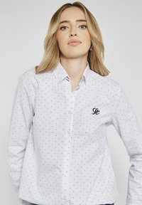 Biała koszula z guzikami, w czerwone i granatowe kropki, z małym granatowym logo po lewej stronie oraz klasycznym kołnierzykiem.