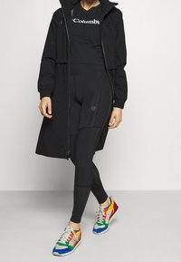 Svart zip-up jacka med huva, svarta åtsittande leggings och flerfärgade sneakers med blå, grön, gul och röd accent.