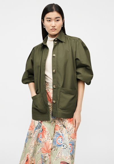 Γυναίκα που φοράει χακί σακάκι με κουμπιά και μεγάλες τσέπες πάνω από κρέμ τοπ και φλοράλ φούστα με παστέλ χρώματα.