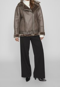 Giacca di pelle marrone con un ampio colletto, chiusura con zip e foderata in pelliccia. Abbinata a pantaloni neri larghi e scarpe nere a punta.