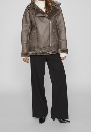 Chaqueta de cuero marrón con un cuello ancho, cierre de cremallera y forro de piel. Combinada con pantalones negros holgados y zapatos negros de punta.