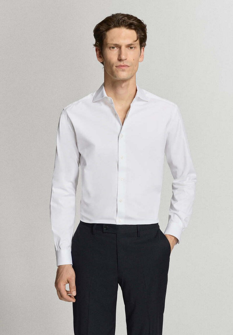Homme portant une chemise blanche cintrée à boutons et un pantalon noir, debout devant un fond clair uni.