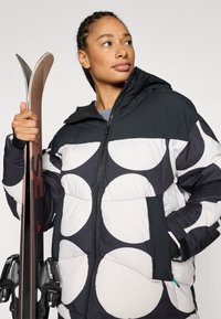 Gewatteerd jack met een zwart-wit cirkelpatroon, voorzien van een ritssluiting aan de voorkant en een capuchon, gedragen door een persoon die ski's vasthoudt.