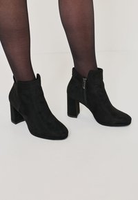 Black suede ankle boots with a block heel, pointed toe, and side zipper detail, showcased with black sheer tights on a white background.