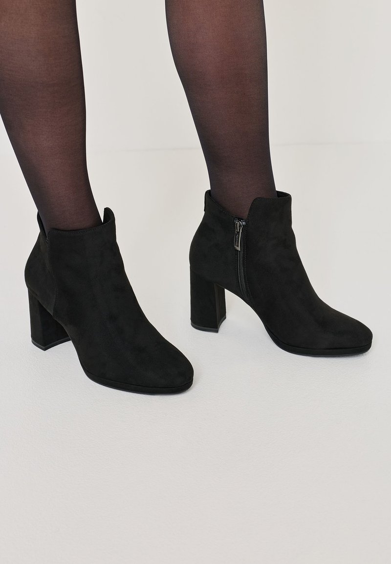 Black suede ankle boots with a block heel, pointed toe, and side zipper detail, showcased with black sheer tights on a white background.