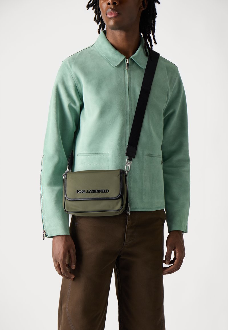 Giacca di suede verde con zip e due tasche, abbinata a pantaloni marroni. Una piccola borsa a tracolla verde oliva presenta lettere nere.