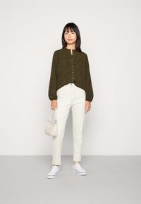 Camisa botonê verde-oliva com colarinho redondo e estampado, calças de denim brancas e sapatilhas brancas; segurando uma pequena bolsa de creme.