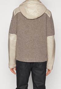 Giacca marrone in misto lana con dettagli in satin beige, cappuccio e texture morbida. Presenta un dettaglio con logo all'orlo e materiali a contrasto sulle maniche.