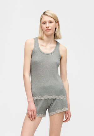 Mladá žena v sivej rebrovanej tielkovej vesta a zodpovedajúcich šortkách s čipkovým lemom, stojaca pred čistým bielym pozadím.