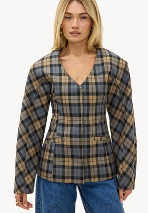 Femme portant un haut à carreaux bleu et beige à col en V avec des manches longues bouffantes et un jean bleu, sur un fond uni.