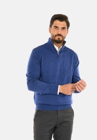 Maglione blu con cerniera realizzato in materiale morbido, caratterizzato da un colletto alto e polsini a costine. Abbinato a pantaloni grigi.