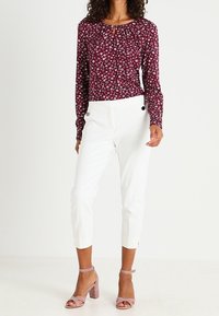 Blusa a maniche lunghe color bordeaux con motivo floreale, pantaloni bianchi su misura e sandali con tacco a blocco rosa. La blusa presenta un collo rotondo e una leggera arricciatura.