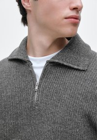 Maglione grigio lavorato a maglia con mezza cerniera, dalla trama a coste e ampio colletto, indossato sopra una camicia bianca. Dettaglio della cerniera argentata sul davanti.