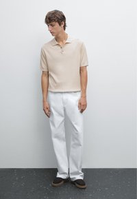 Beige ribstrikket poloshirt med krave, kombineret med løse hvide bukser og brune slip-on sko, stående mod en neutral baggrund.