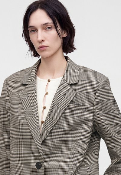 Jeune personne aux cheveux courts et foncés portant un blazer beige et marron à carreaux sur un haut blanc avec des boutons en bois.