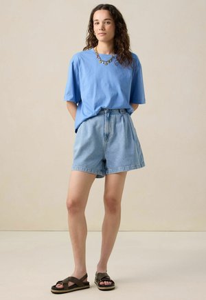 Lichtblauwe katoenen T-shirt met korte mouwen, gecombineerd met hooggetailleerde denim shorts en bruine sandalen met bandjes. Een eenvoudige ketting accentueert de look.