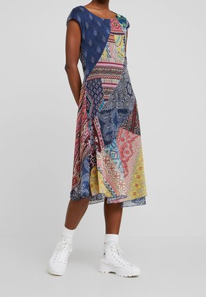 Kvinde iført en farverig patchwork midi-kjole med kappelignende ærmer, kombineret med hvide høje sneakers og sokker, stående foran en grå baggrund.