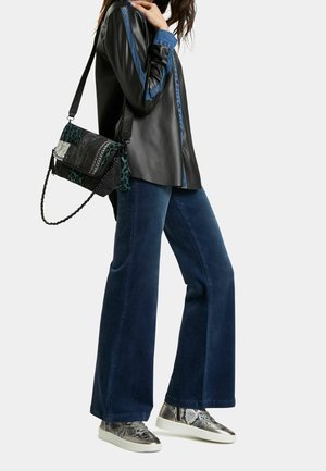 Femme portant une chemise en cuir noire avec des bordures bleues, un jean large bleu foncé, des baskets métalliques à imprimé serpent, et portant un sac à épaule noir et vert à motifs.