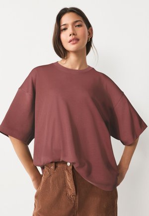 Femeie cu păr drept și închis la culoare poartă un top oversized mauve cu mâneci scurte și pantaloni maro de catifea, pozând cu mâinile în buzunare pe fundal alb.