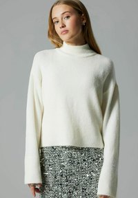 Valge, pehme kootud sviiter kõrge kraega, langenud õlgade ja laiate varrukatega, kombineeritud särava hõbedase sädelevate sequin-seelikuga.