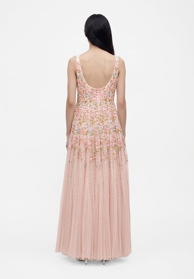 Femme aux longs cheveux noirs portant une robe rose sans manches avec broderies florales et un dos échancré, debout devant un fond blanc uni.