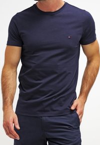 Uomo che indossa una maglietta a maniche corte blu navy attillata con un piccolo logo rosso, bianco e blu sul lato sinistro del petto e pantaloni blu navy.