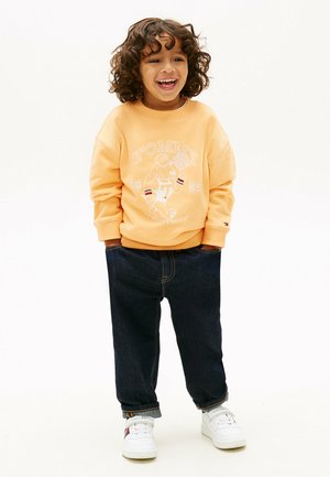 Smilende barn med krøllet hår iført en ferskenfarvet sweatshirt med et hundebasketballmotiv, mørke bukser med opsmøg og hvide sneakers.