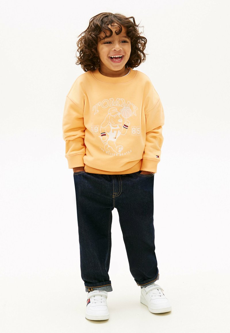 Enfant souriant aux cheveux bouclés portant un sweat-shirt pêche avec un motif de chien jouant au basketball, un jean foncé retroussé et des baskets blanches.