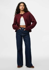 Bordeaux gebreid vest met een knoopsluiting, cropped lengte en brede mouwen, gecombineerd met hoogwaardige donkerblauwe jeans.