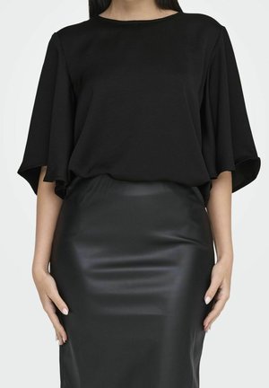 Vrouw draagt een losse zwarte top met wijde mouwen en een nauwsluitende zwarte leren rok, staand met handen ontspannen langs het lichaam.