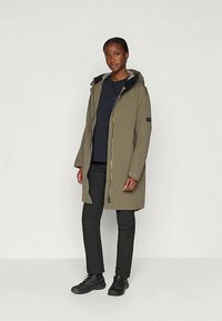 Olijfgroene jas met een rits aan de voorkant, capuchon en zwarte accenten. Gecombineerd met een marineblauw shirt, zwarte broek en donkere schoenen. Gladde textuur.