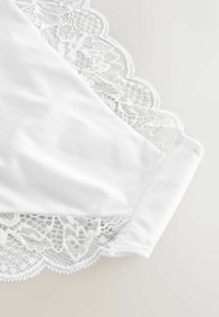 Tissu blanc avec bordure en dentelle festonnée présentant des motifs floraux et des détails en mesh, exposé sur un fond clair.